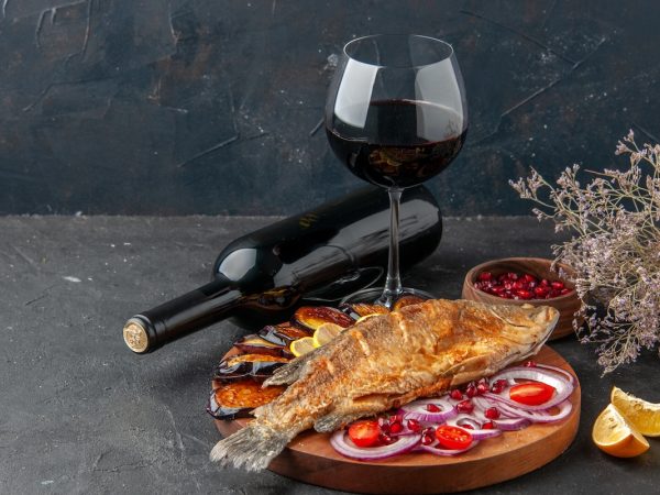 front view fish fry fried aubergines cut onion on wood serving board wine bottle and glass on dark background